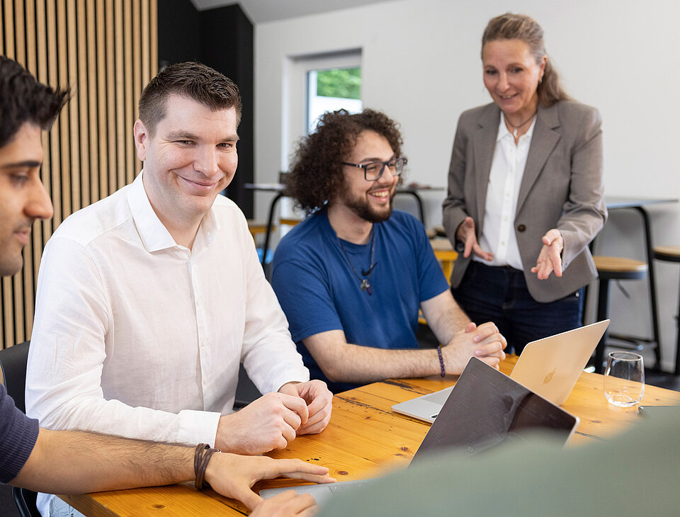 Das Team von Rollout Solutions arbeitet gemeinsam an einem großen Holztisch, ein Mann im weißen Hemd lächelt in die Kamera