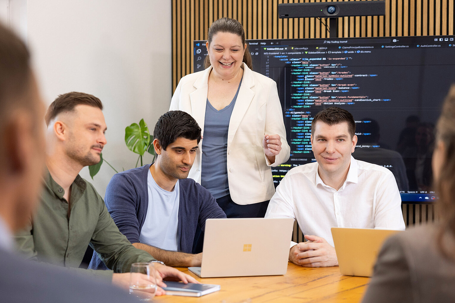 Männliche und weibliche Teammitglieder von Rollout Solutions arbeiten gemeinsam am Laptop, im Hintergrund ein großer Bildschirm mit Codezeilen
