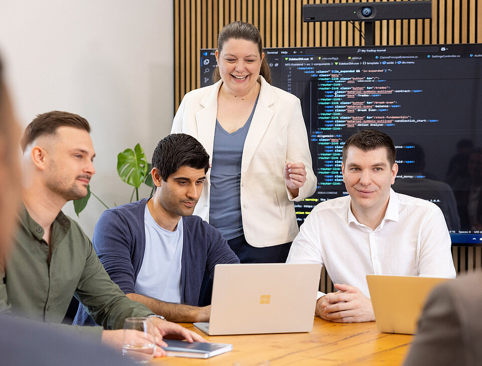 Männliche und weibliche Teammitglieder von Rollout Solutions arbeiten gemeinsam am Laptop, im Hintergrund ein großer Bildschirm mit Codezeilen