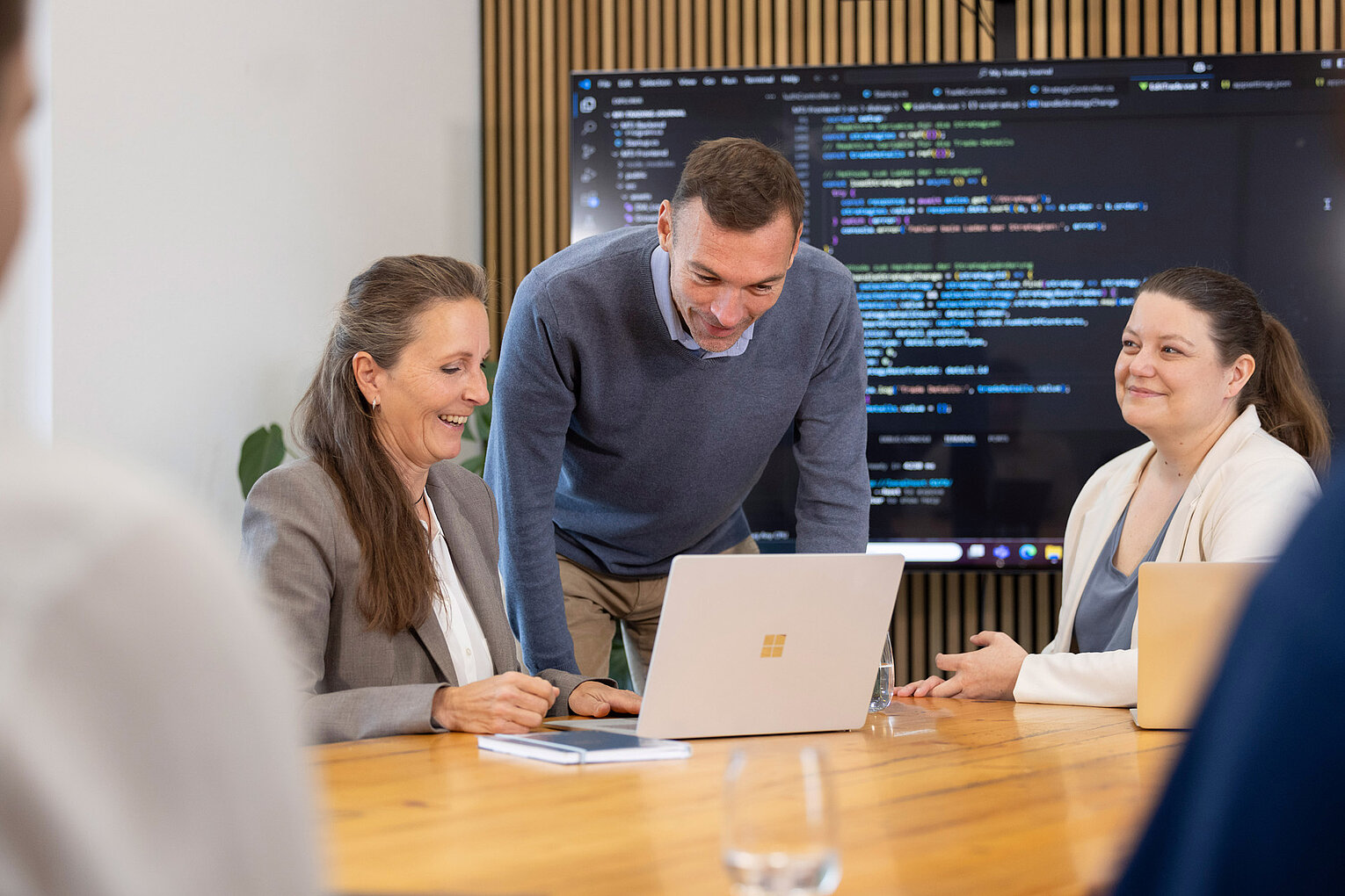 Männliche und weibliche Mitarbeiter von Rollout Solutions blicken gemeinsam auf einen Laptop, im Hintergrund ein Bildschirm mit Code