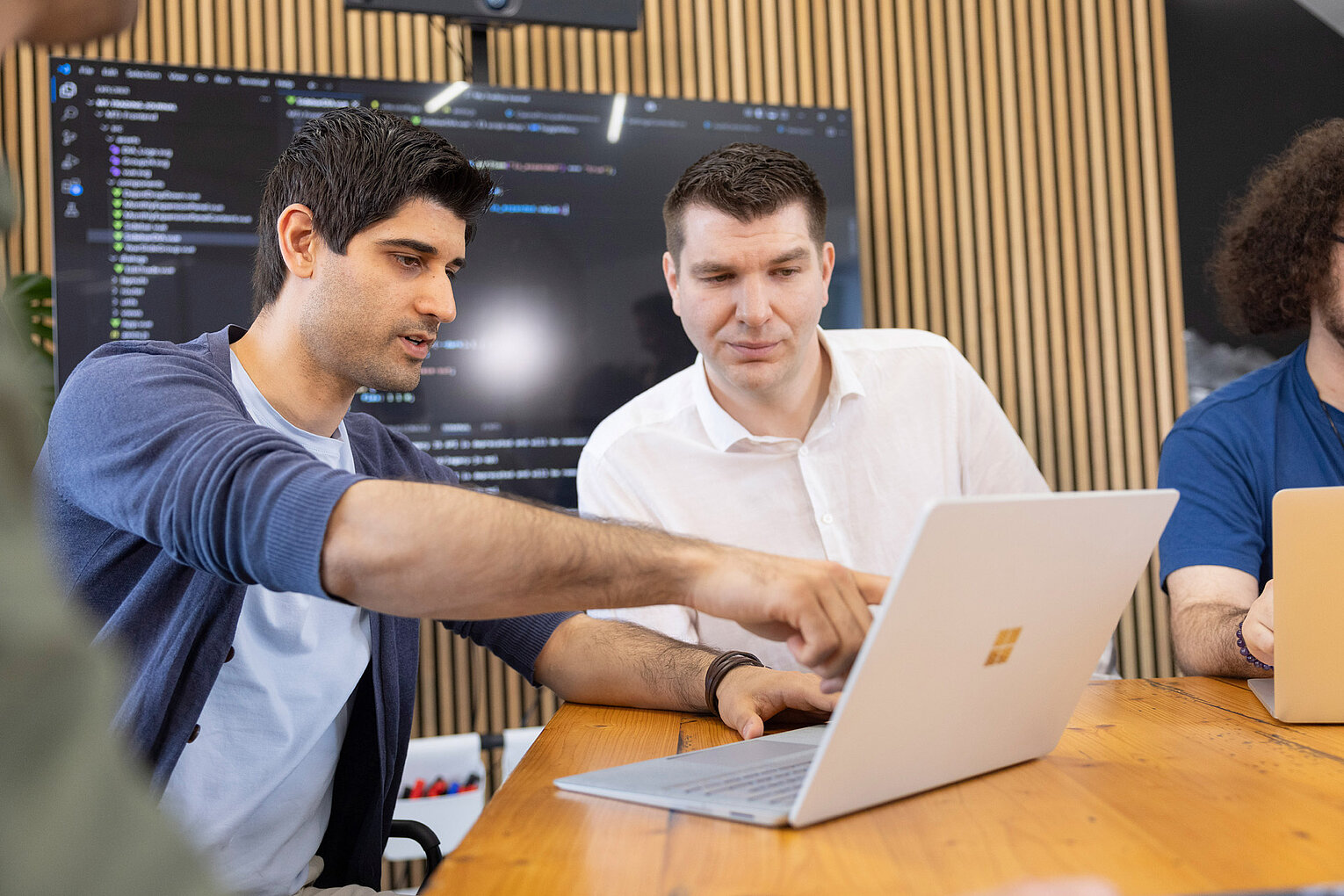 Ein Mitarbeiter von Rollout Solutions zeigt einem Kollegen etwas auf dem Display eines Laptops, im Hintergrund ein Bildschirm mit Code