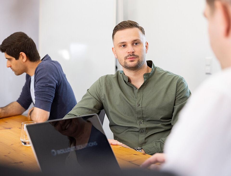 Tomislav Tomic von Rollout Solutions im Gespräch mit einem Arbeitskollegen an einem Tisch, im Vordergrund ein Laptop