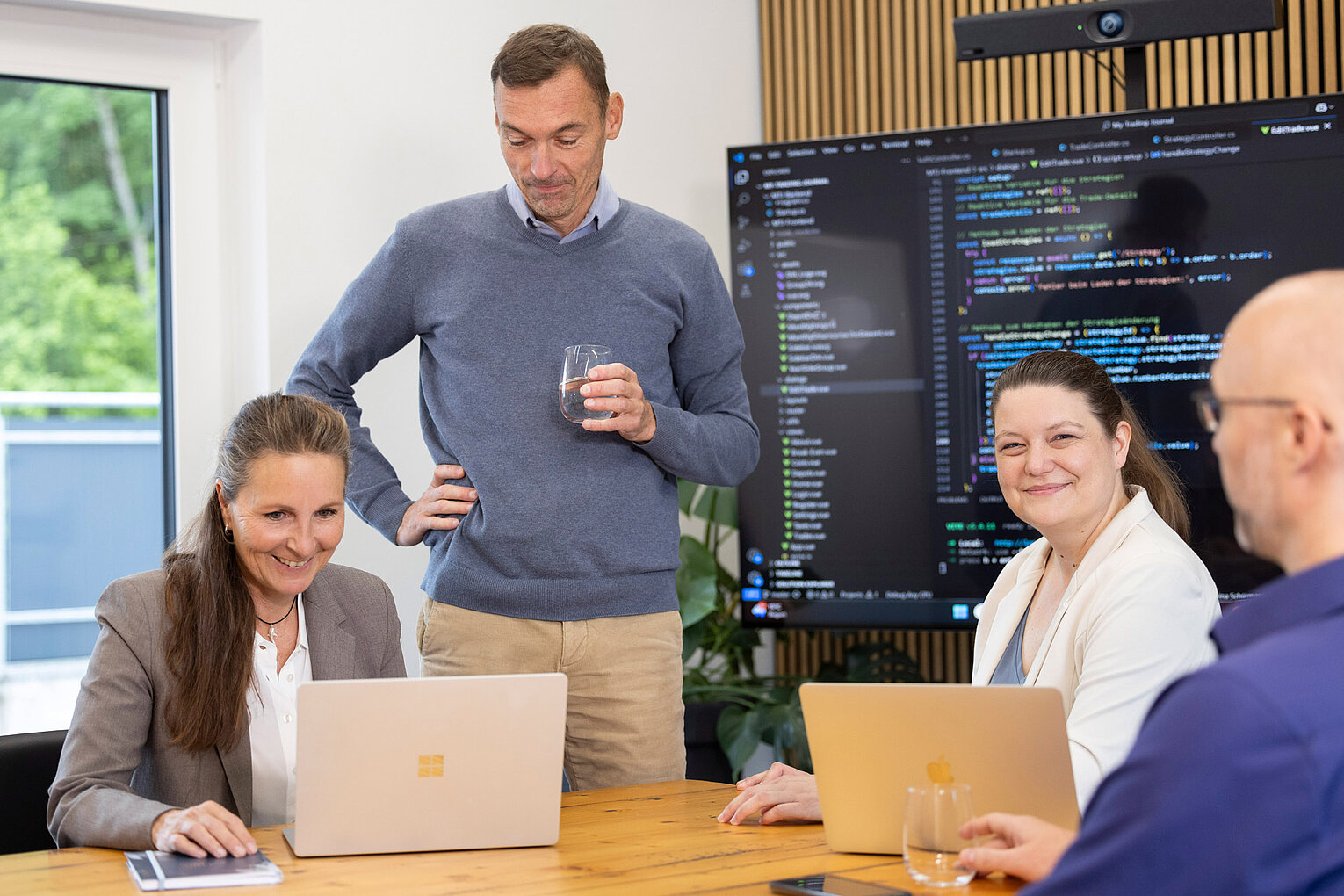Bernd Stiller von Rollout Solutions steht hinter den Geschäftsführerinnen und schaut auf das Display eines Laptops, im Hintergrund ein großer Bildschirm mit Code