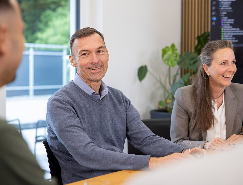 Ein Mitarbeiter von Rollout Solutions unterhält sich am Tisch mit Kollegen, im Hintergrund eine lachende Frau